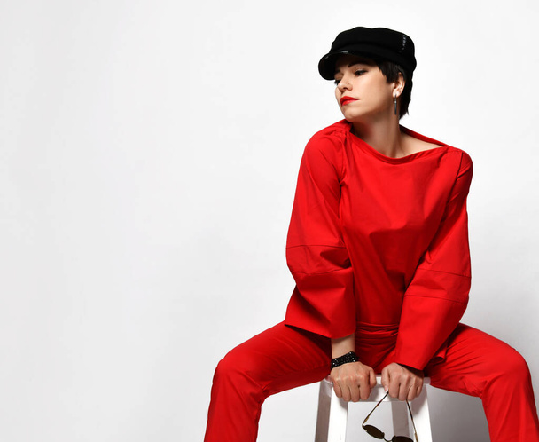 Adult pretty sensual short haired brunette woman in stylish casual red pantsuit and black cap sits on chair - Foto, imagen