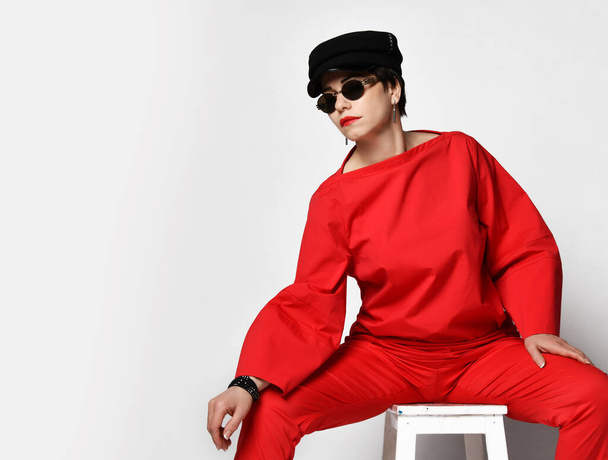 Mujer morena de pelo corto, artista, cantante en traje pantalón rojo de alta costura, gafas de sol y gorra negra se sienta en la silla - Foto, imagen