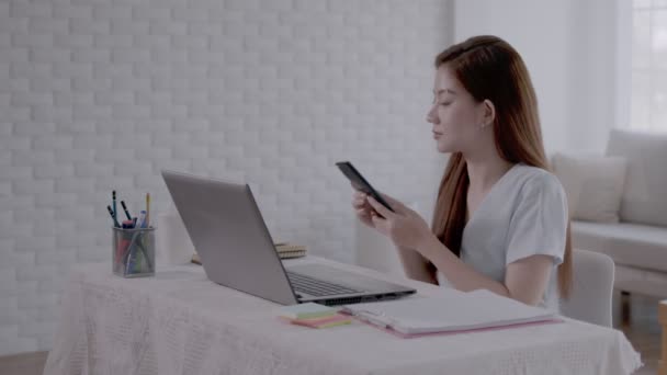 Деловая азиатская женщина разговаривает по телефону и делает заметки на ноутбуке с работой из дома. - Кадры, видео