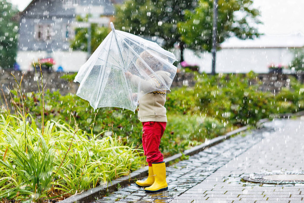 Vaalea pikkupoika kävelee sateenvarjon kanssa ulkona sadepäivänä. Esikoululainen hauskaa ja yllään värikäs vedenpitävä vaatteita ja sadekumia kumisaappaat. Ulkoilu huonona sääpäivänä - Valokuva, kuva