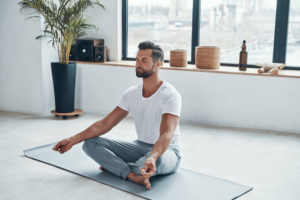 Totally relaxed young man doing yoga while sitting in lotus position at home - Photo, image