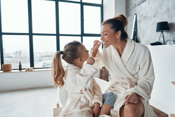 Mother and daughter in bathrobes smiling and cleaning teeth while doing morning routine - Foto, afbeelding