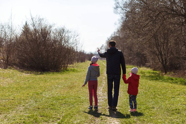 Νεαρός πατέρας με δύο κόρες που περπατούν στη φύση. Ένας άντρας με δύο παιδιά περπατάει στο μονοπάτι, από πίσω. Ο μπαμπάς κρατάει τα παιδιά από τα χέρια.. - Φωτογραφία, εικόνα