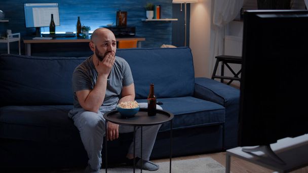 Сосредоточенный мужчина смотрит драматический фильм, плачет, сидя на диване и поедая попкорн - Фото, изображение
