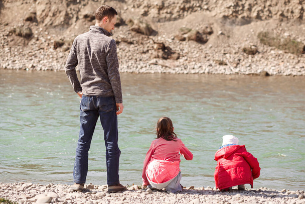 Papà e figlie stanno sulla riva rocciosa del fiume. Un uomo con dei bambini seduto vicino al fiume. Foto di famiglia sulla spiaggia del fiume. - Foto, immagini