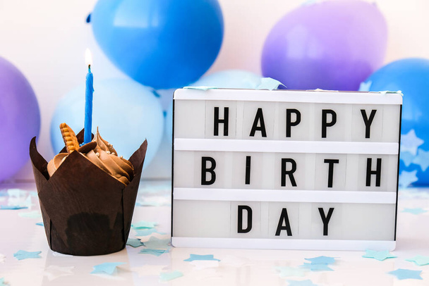 テキスト付きのライトボックスハッピー誕生日とチョコレートカップケーキキャンドル抽象的に焦点を当てた休日のためのぼやけたお祝いの背景。青い風船とコンフェッティ、誕生日ケーキ。グリーティングカードボーイコンセプト - 写真・画像