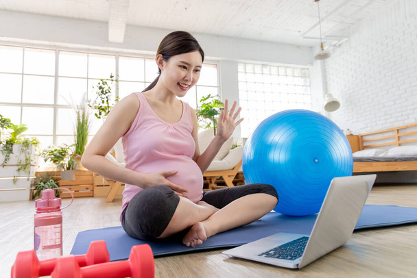 Asian young pregnant woman using laptop to have online exercise class with pilates ball and dumbbells in the living room - saying hi - Фото, зображення