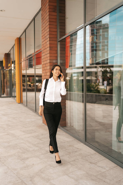 Femme avec backpak d'affaires marchant tout en parlant au téléphone près d'un immeuble d'affaires - Photo, image