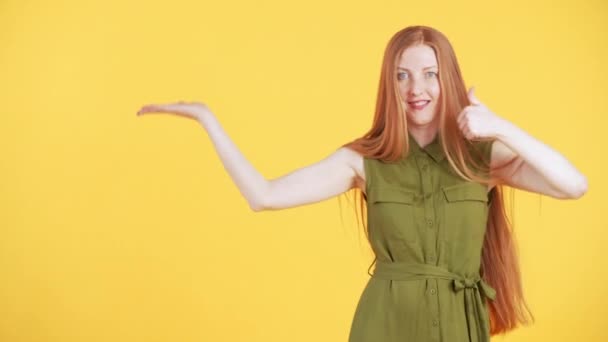 Jolie femme avec des taches de rousseur et de longs cheveux roux regarde dans la caméra et danse d'une manière drôle avec ses pouces vers le haut sur fond jaune - Séquence, vidéo