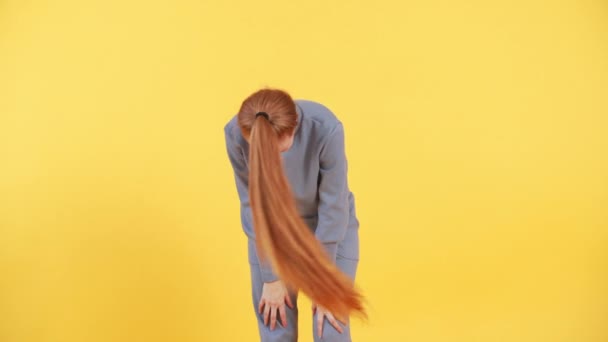 Woman with long ginger hair is twirling her ponytail in a funny way on yellow background in studio - Footage, Video