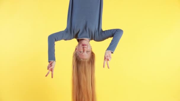 Woman with long ginger hair is hanging upside down, looking into the camera and making funny gestures. Yellow background - Footage, Video