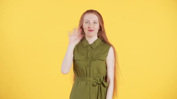 Young woman with freckles on face and long ginger hair is looking into the camera, smiling and waving for a few times. Yellow background - Footage, Video