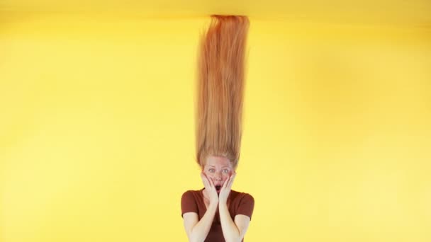 Kobieta z piegami i długimi rudymi włosami nad głową robi zaskakującą minę. Kobieta z długimi włosami wisi do góry nogami. Żółte tło - Materiał filmowy, wideo