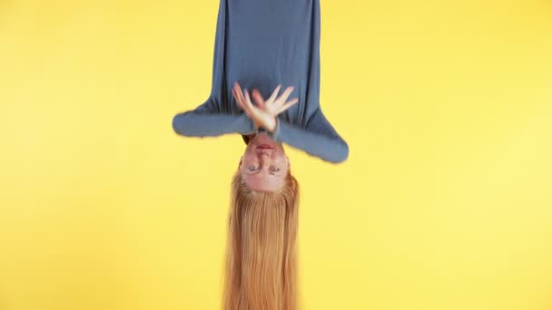 Donna con lunghi capelli rossi è appesa a testa in giù, guardando nella fotocamera e agitando. sfondo giallo - Filmati, video