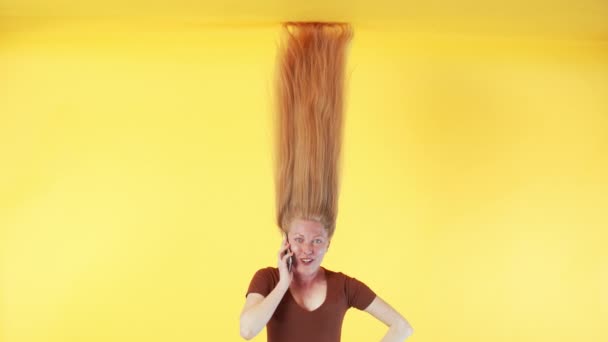 Woman with freckles and with long ginger hair above her head is talking on smartphone and smiling. Woman with long hair is hanging upside down. Yellow background - Footage, Video