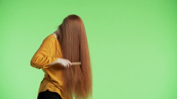 Молодая привлекательная женщина расчесывает длинные рыжие волосы, поднимает голову и смотрит в камеру с улыбкой. Зеленый фон - Кадры, видео