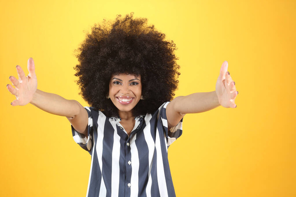 Молодая брюнетка афро женщина и случайный свитер на изолированном желтом фоне глядя на камеру улыбается с распростертыми объятиями для объятий. - Фото, изображение