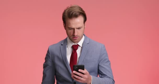 hombre de negocios preocupado en traje elegante con teléfono en las manos leyendo malas noticias, decepcionándose, tocando la barbilla y asintiendo de una manera negativa en el fondo rosa en el estudio - Metraje, vídeo