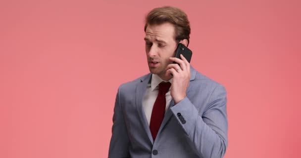elegantní mladý muž v modrém obleku uvolňující kravatu, zatímco mluví po telefonu, přikyvuje a dělá obličeje na růžovém pozadí ve studiu - Záběry, video