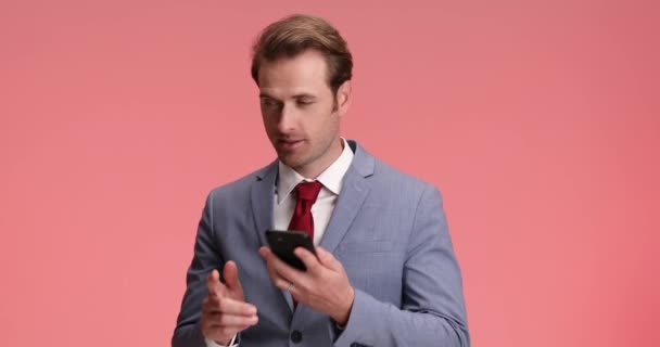 hombre de negocios feliz en traje elegante cogiendo el teléfono, hablando y teniendo una buena conversación y apuntando con entusiasmo el dedo sobre el fondo rosa - Imágenes, Vídeo