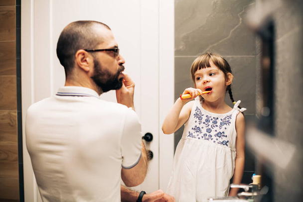 Bearded father and his little daughter brushing teeth in a bathroom and looking at each other. Man teaching his daughter how to brush teeth. - Zdjęcie, obraz