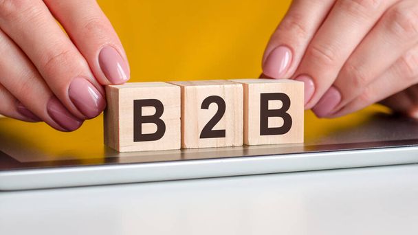 Frauenhände legen einen Holzblock mit den Buchstaben b2b auf die schwarze Oberfläche des Notizblocks. Kann für Business, Marketing, Bildung, Konzept verwendet werden. Gelber Hintergrund. b2b - business to business - Foto, Bild