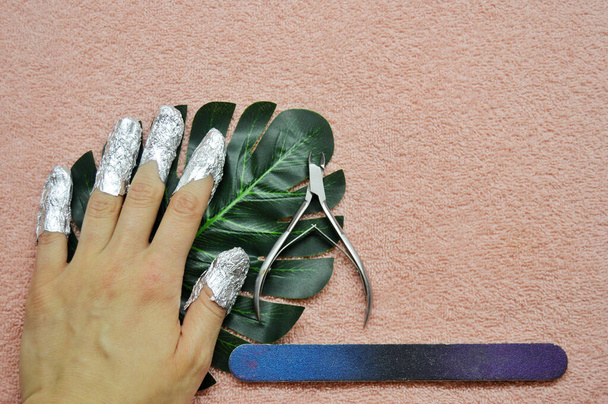 Removing shellac from nails. fingers with foil, wire cutters and nail file on a green leaf of a palm tree on a pink background. Close-up hand. Front view. - Foto, Imagem