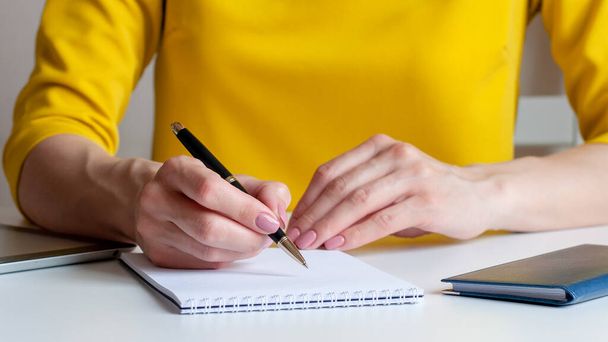 Κοντινή εικόνα μιας γυναίκας που γράφει σε ένα λευκό κενό σημειωματάριο. Κίτρινο φόντο, επιλεκτική εστίαση. Μπορεί να χρησιμοποιηθεί για επιχειρήσεις, μάρκετινγκ, εκπαίδευση, οικονομική έννοια. - Φωτογραφία, εικόνα