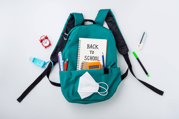Sac à dos turquoise avec fournitures scolaires et masque pour enfants sur fond blanc, vue de dessus - Photo, image