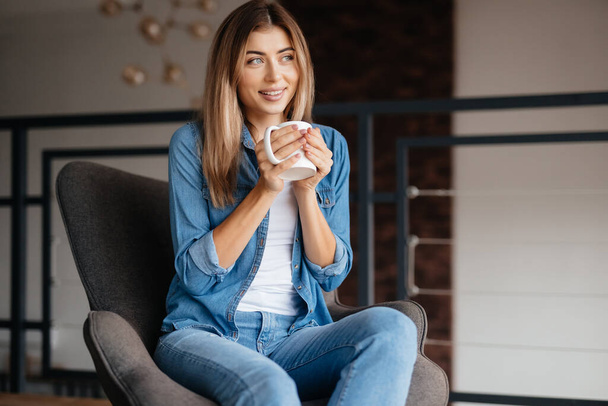 Fair-haired young woman looking dreamy while holding cup of coffee and looking to the side - 写真・画像