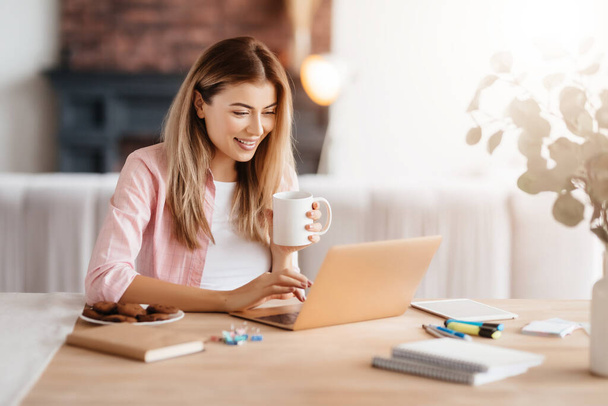 Delighted young lady with coffee cup sitting at desk and looking at laptop screen - Фото, зображення