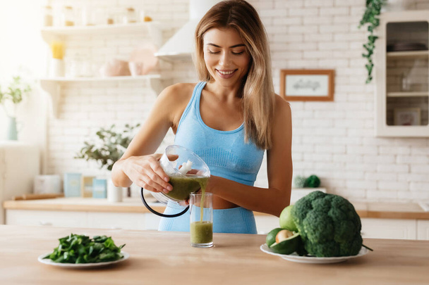Gladsome young woman smiling while puring freshly-made green smoothie into a tall glass - Foto, Bild