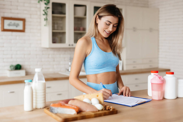 Attractive lady taking notes and standing near kitchen table with meat, dairy products and food supplements on it - Фото, зображення