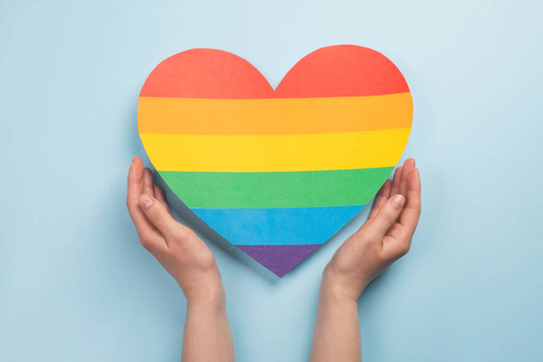 Frauenhände halten Herzen in den Farben des Regenbogens auf hellblauem Hintergrund. LGBT-Konzept, Pride Monat. Ansicht von oben - Foto, Bild
