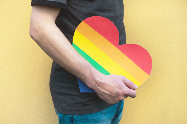 Mann mit Herz in den Farben des Regenbogens auf gelbem Hintergrund. LGBT-Rechte, Pride Monat, Toleranz und Liebeskonzept. Nahaufnahme - Foto, Bild
