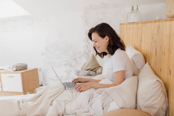 Happy beautiful woman working on a laptop while sitting on a bed in a bright modern house. - Valokuva, kuva