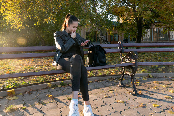 Студент сидить на лавці в парку і дивиться в смартфон. Осінній сонячний день
 - Фото, зображення