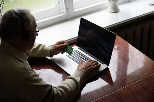 Lächelnder älterer Mann mit Kopfhörer winkt mit Videoanruf auf Laptop in die Kamera, glücklicher älterer Mann mit Kopfhörer sitzt auf Couch zu Hause und spricht mit modernen Technologien und drahtloser Verbindung - Foto, Bild