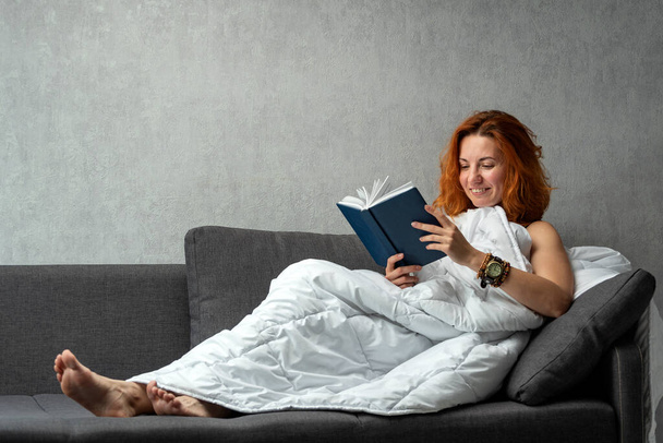 Menina ruiva jovem no sofá sob cobertor lê livro. Feliz repouso mulher. - Foto, Imagem