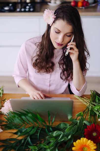 Jeune fleuriste regardant ordinateur portable et répondre à un appel téléphonique - Photo, image