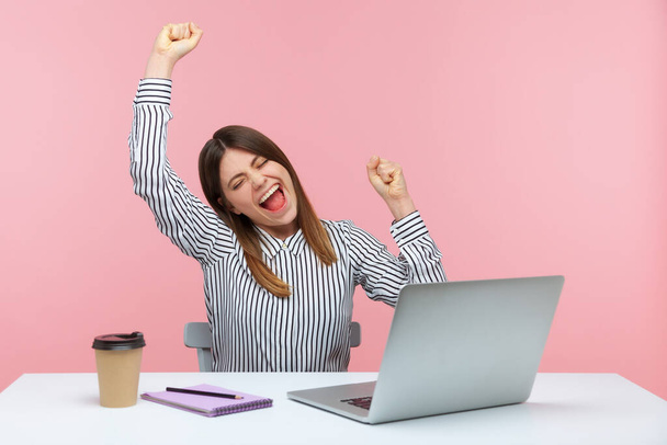 Happy excited young woman office worker raising hands and screaming for joy, celebrating successful business online, sitting at workplace. Indoor studio shot isolated on pink background - Foto, Bild