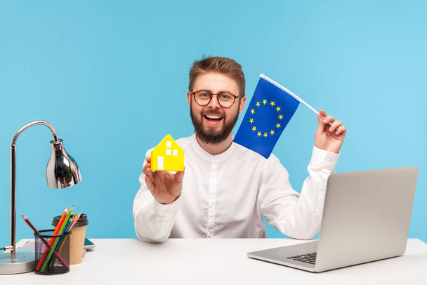 Smiling positive man real estate agency worker in eyeglasses holding paper toy house and flag of europe union, registration of visas and passports. Indoor studio shot isolated on blue background - Foto, Imagem