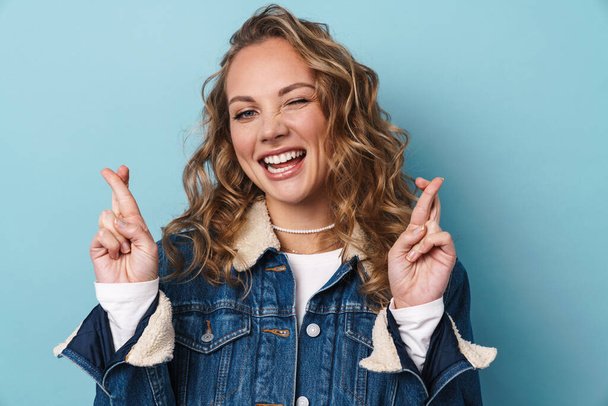Blonde happy woman winking and holding fingers crossed for good luck isolated over blue wall - Photo, image