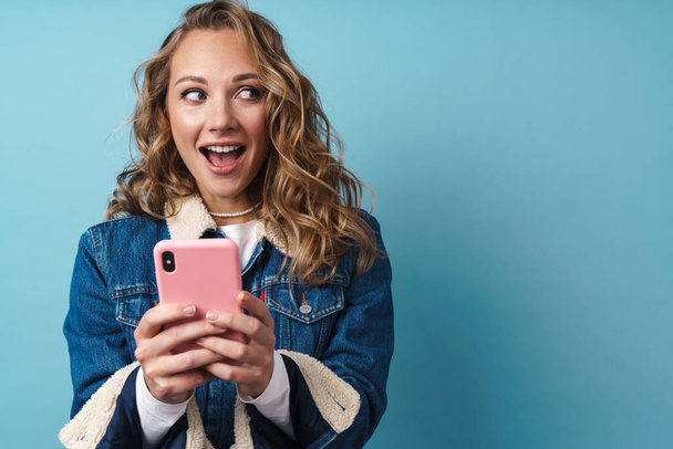 Blonde ενθουσιασμένος γυναίκα χρησιμοποιώντας το κινητό τηλέφωνο και χαμογελώντας απομονωμένη πάνω από το μπλε τοίχο - Φωτογραφία, εικόνα