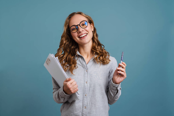 Happy girl in eyeglasses smiling while posing with papers isolated over blue background - Photo, Image