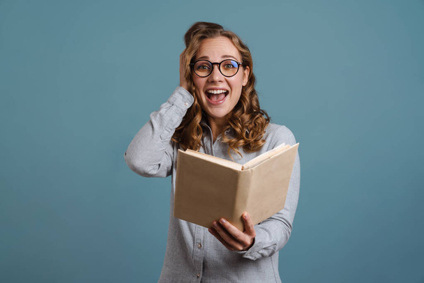 Excited beautiful girl in eyeglasses reading book isolated over blue background - Фото, зображення