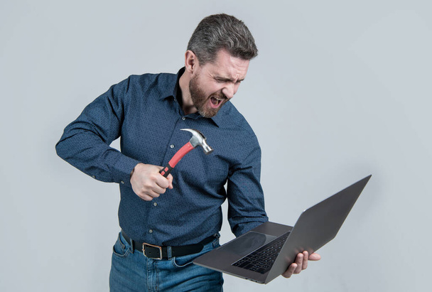 Εξοργισμένος άνθρωπος διάλειμμα laptop χτύπημα σημειωματάριο με σφυρί γκρι φόντο, οργή υπολογιστή. - Φωτογραφία, εικόνα