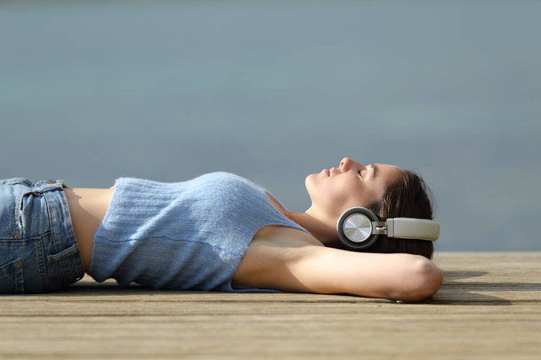 Profile of a woman wearing wireless headphones relaxing listening to music in a lake pier - Fotoğraf, Görsel