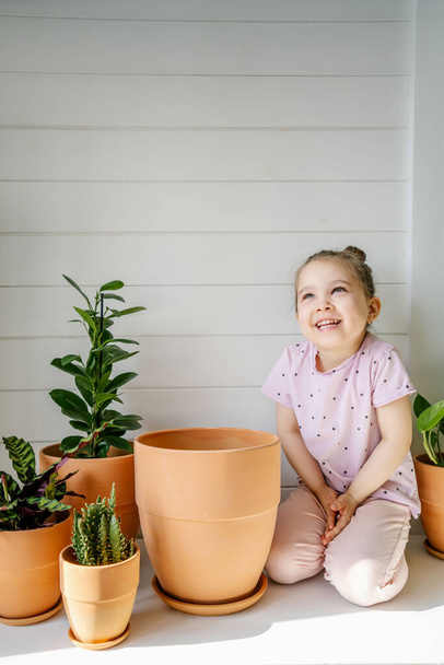 A girl sits on a white dresser between flower pots. - Photo, Image