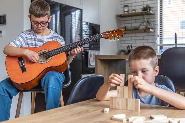 Pieni poika soittaa kitaraa, ja hänen veljensä rakentaa tornin, jossa on puisia kuutioita kotona pöydässä.. - Valokuva, kuva